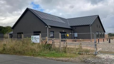 Large grey building behind temporary fencing. Solar panels are on the roof. 