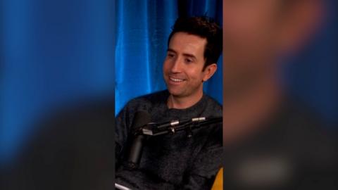A man, Nick Grimshaw, smiling in a podcast studio, with a blue curtain behind.