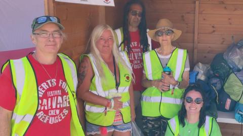 Two men and three women all wearing high vis jackets by a shed which is full of tents wrapped up in their bags. The men are wearing red T-shirts and one of the women is wearing a green T-shirt. There is a sign saying "Tent Donation Point" at the top of the shed.