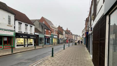 A picture of a street in Atherstone, Warwickshire. A road stretches into the distance with terraced shops on either side. On either side of the road are brick pavements and dark green bollards.