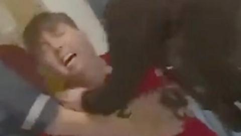 Jamie Varley wears a red t-shirt and is kneeling on the floor, he is surrounded by a police officer and a nurse