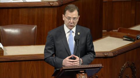 A man in a suit stands at a podium holding his hands together