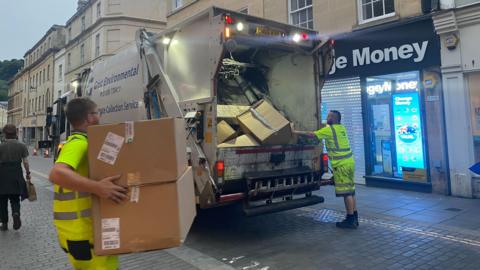Two waste removal workers putting boxes into the back of a waste truck. They are wearing hi-vis uniforms.