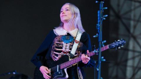 Phoebe Bridgers performs on stage holding a black guitar. She is a young woman with short, grey-coloured hair. She is wearing a black dress with a white colour and a decorated ribcage on the front of it.