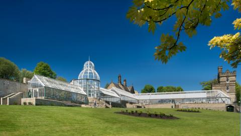 A white framed Victorian-style greenhouse complex with a green lawn in front of it and a turreted building in stone to the right-hand side in the background.  