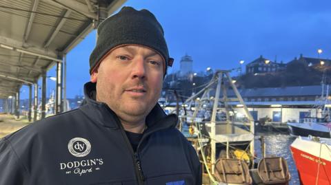 Colin Graham a fisherman, dressed in a navy jumper and wooly hat stands in front of docked boats at the North Shields fish quay at the evening. 