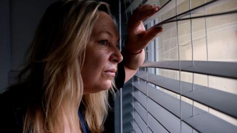 Catherine stares out of the window through her blinds