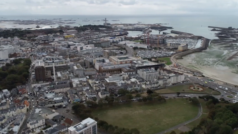 Aerial view of St Helier with buildings and roads close together, a large park bottom right and the beach to the right, the sea behind and grey skies.