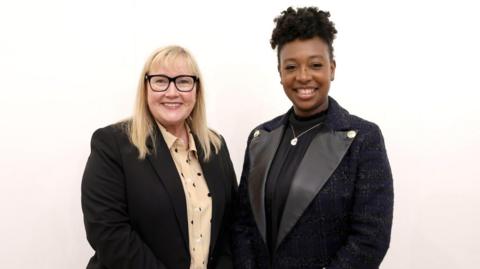 Newcastle City Council leader Karen Kilgour and renowned musician YolanDa Brown stand side-by-side against a white background. Karen Kilgour wears a black suit jacket and a cream and black polka dot shirt. YolanDa Brown wears a black suit jacket and shirt. They are smiling at the camera.