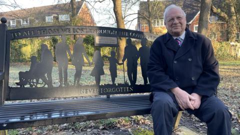 An elderly man with white hair sitting on a black bench which says "To all those we lost through the Covid-19 pandemic. Never Forgotten" on it. He is wearing a dark felt coat with a pink and blue tie underneath.