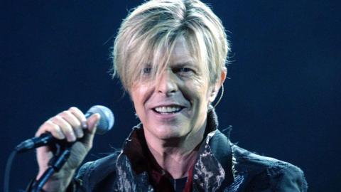 Head and shoulders of David Bowie looking at the camera while holding a microphone on stage. The background is very dark and Bowie has a long blonde fringe and is wearing a raggy-looking jacket