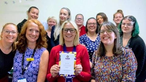 A group of women all smiling in a room while a woman with blond shoulder length hair, glasses and a red top is smiling holding a certificate that has a star on it and says "Cavell" in purple print.