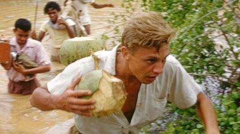 An image of a young Attenborough crossing a body of water. Three men can be seen in the background behind him.