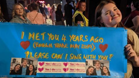 A young girl with brown hair smiling holding a sign that says 'Can I sing with you'