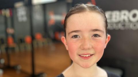 Ten year old Martha is looking at the camera, she is fair skinned with freckles and her hair is in a tight bun. Behind her is a dance studio. 