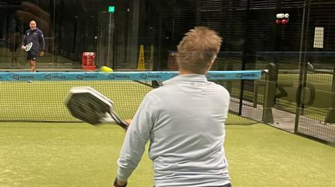 A general view of two people playing padel at an indoor court. One person, with short blond hair and wearing a grey sweater, is in action having just hit the ball. They are pictured from behind and the ball is travelling towards a man in blue shorts and sweater preparing to received the ball.