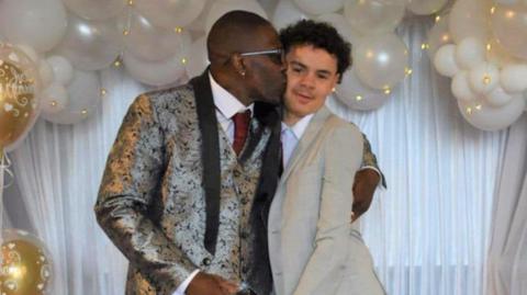 A father kisses his son on the cheek - both wearing suits, surrounded by balloons at a wedding.
