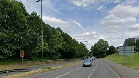 A Google Street View image showing the dual carriageway of the A25 through Guildford. There are three lanes of traffic on the near side and two on the other, with a grass verge and trees at the side of the road.