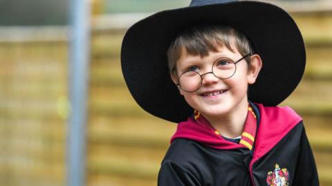 A boy dressed as Harry Potter, with round glasses, brown hair, a black wizard hat and black and red gown with a logo. He is smiling.