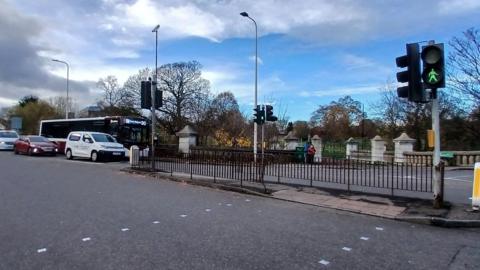 A street view of a pedestrian crossing on Edinburgh's Gorgie Road in front of Saughton Park while the green man is showing. 