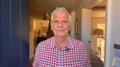 Peter Robinson - a man in a red and white shirt. He is tanned and has white hair. He has piercing blue eyes. He is standing in the doorway of his home that looks like a cottage.