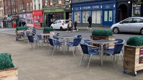 A city centre street with a seating area on the pavement. There are shops lining the street and people and cars pictured in the background.