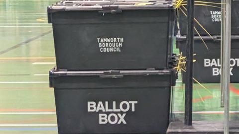 A stack of black ballot boxes marked “Tamworth Borough Council” and “Ballot Box” inside a large indoor hall with a green floor.
