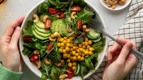 An above view of a white bowl which has a salad in, containing cucumber, avocado, tomatoes and leaves.  A person is holding a fork with their right hand.