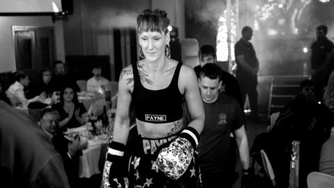 Lucy Payne in her boxing clothes and gloves standing in front of people eating at tables. The picture is black and white.