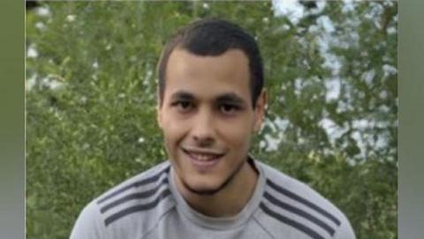 Kurtis Brook smiling at the camera. It is a head and shoulders shot of him - he has short dark hair and dark short stubble. He is wearing a grey sweatshirt with stripes on the shoulders. 