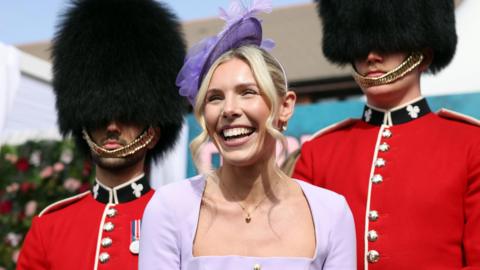 A woman in a lilac dress and fascinator smiles in front of two guardsmen wearing full parade uniform of black busby hats and red jackets with black collars