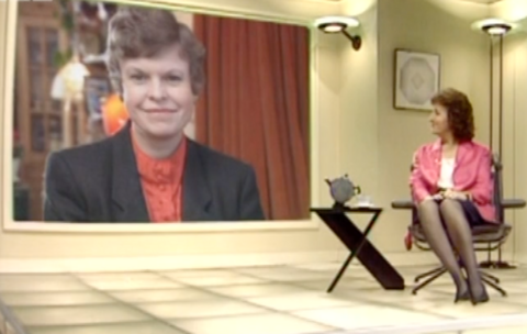 Judith Hann sitting in a chair in the set, with Christine McNulty on a large screen behind her.