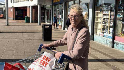 Lorraine Monaghan, a mature woman with long blonde hair with a fringe and glasses holds onto her shopping trolley outside the Braes shops. The trolley is full of bags from different shops. She wears a dusky pink jacket.