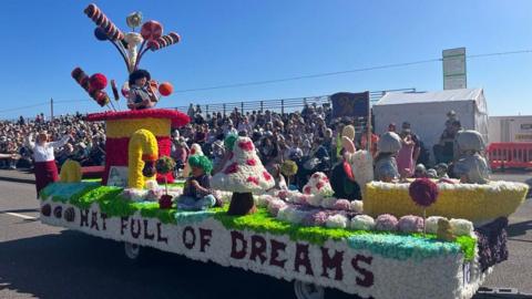 A colorful parade float decorated with large mushrooms, candy canes, and whimsical characters in costumes. The float features the text 'HAT FULL OF DREAMS' on its side. Spectators are seated in bleachers in the background, watching the parade.