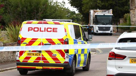 The back of a police vehicle parked in a road, with police tape in front and the white bin lorry in the distance behind it