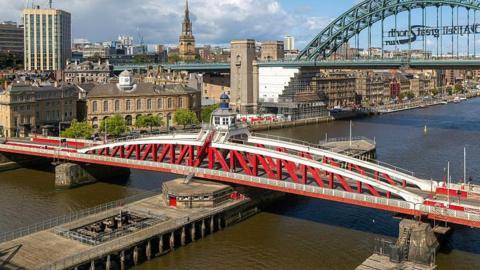 The Swing Bridge is red and white arched bridge and spans the River Tyne. There is a wooden platform underneath which the bridge is designed to swing on to. The Tyne Bridge and a city scape appears in the background. 