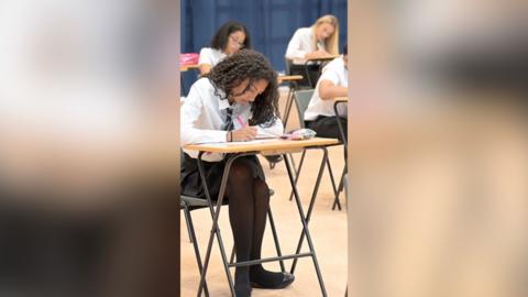 Teenagers sit at desks writing in an exam hall.