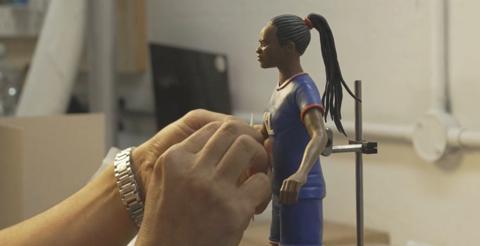 Hands working on a clay model of a female footballer dressed in a blue kit.