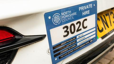 A North Yorkshire Council license plaque for a taxi affixed to a white electric car.
