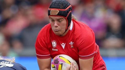 Carys Phillips, wearing a black scrum cap with red trim, carries the ball while in action for Wales