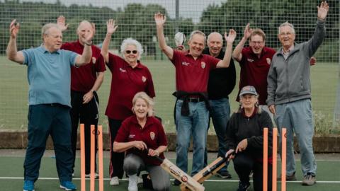 Group of older adults standing on an outdoor court, with several people standing behind a set of bright orange stumps with their hands raised in the air and two others kneeling in front holding cricket bats. There is a fenced sports pitch and greenery in the background.