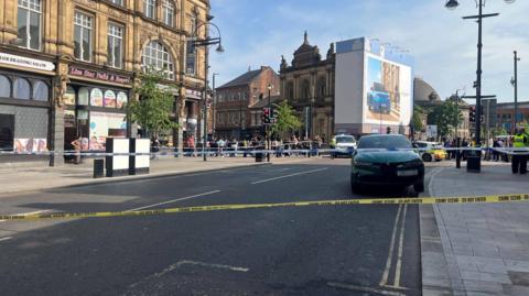 A road with a yellow police cordon around it. There is a car in the middle of the road with police cars and officers in the background. There are shops surrounding the road and crowds of people on the roadside.