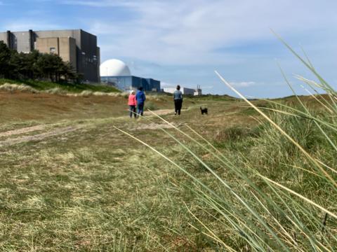 White dome of Sizewell B and other buildings in the background. In the foreground people walk a dog along a footpath across dunes. 