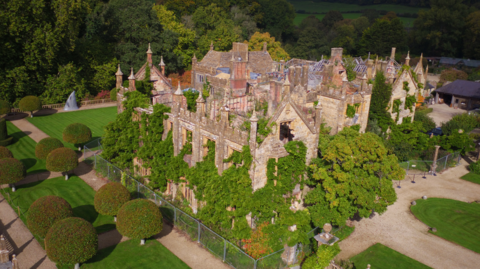 An aerial view of Parnham House. Trees and shrubs are growing out through windows as well as up through the interior of the turreted building, which has no roof in places. By contrast, the surrounding parkland is immaculately landscaped.