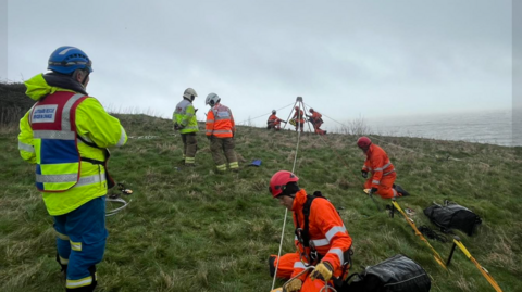 Coastguard rescue teams in orange and yellow overalls standing at the top of a cliff working with a rope technican to rescue a dog.
