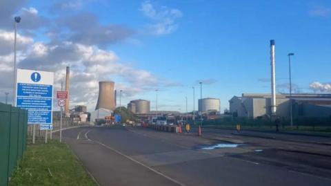 A still of the British Steel plant in Scunthorpe, including cooling towers and large metal buildings. There are safety warning signs at the entrance.