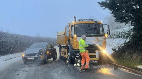 The aftermath of a car crash in the snow. A silver hatchback car has collided with a yellow gritter lorry and the front near side is smashed.