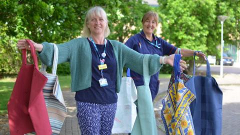 Two nurses stand in a car park in the sun, with large fabric bags hanging off of their arms. They are smiling and the car park is surrounded by trees. It is sunny but they are standing in the shade.