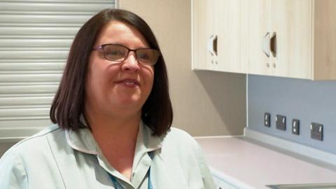 A woman with dark brown hair, glasses and a light green nurse's uniform looks to the right hand side of the camera. Behind her is a work surface with several plug sockets in the wall under four cupboards.