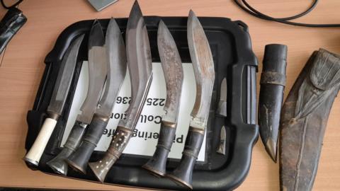 Five large knives and two smaller blades on top of the lid of a plastic box, on a desk in an office.